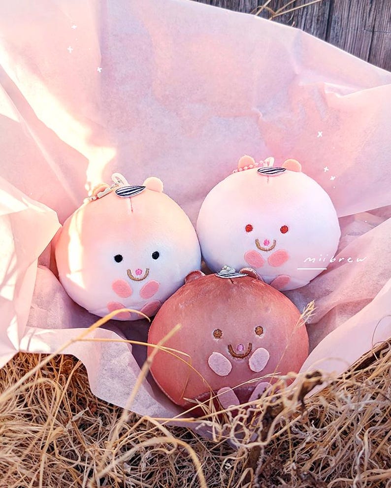 Op de afbeelding: Drie pluche, ronde, hamsterachtige speeltjes in roze en bruine tinten, genesteld in een met roze papier gevoerde mand. Elk speeltje heeft een lachend gezicht met geborduurde details en een kleine hoed. De achtergrond bevat droog gras.