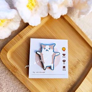 May include: A white and brown cat with blue eyes is depicted on a metal pin. The pin is on a white card with the text "miibrow by miibrow" and a few symbols.