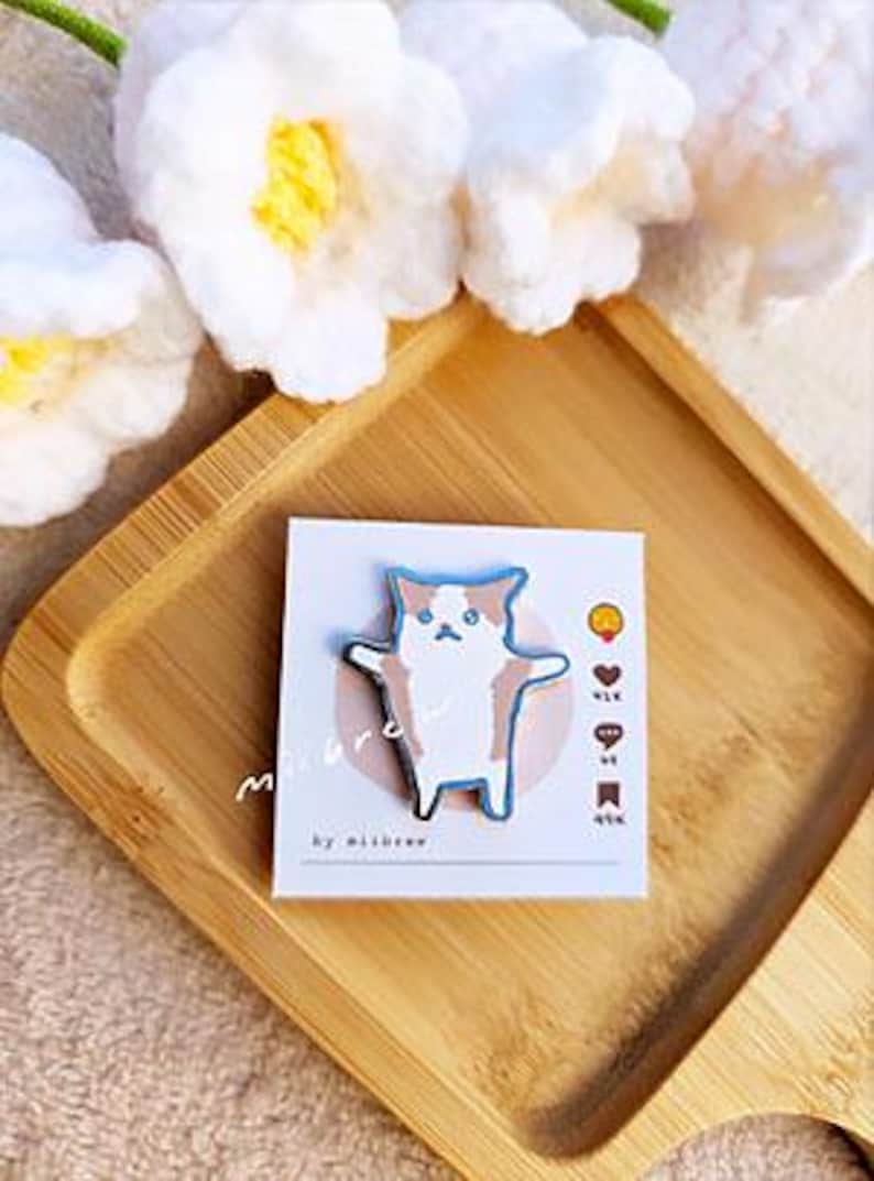 May include: A white and brown cat with blue eyes is depicted on a metal pin. The pin is on a white card with the text "miibrow by miibrow" and a few symbols.