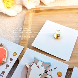 May include: Three enamel pins featuring cartoon cats. The pins are on white cards with text that reads "Locking clutch you won't lose your pins again! pull the top part to unlock" and "milbrew".