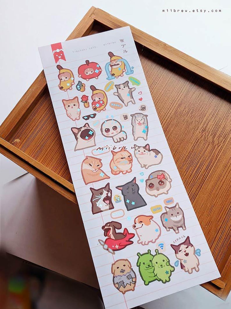 May include: A sheet of stickers featuring various cartoon animals and food items. The stickers include cats, dogs, bananas, and apples, all with cute expressions. The stickers are on a lined paper background.