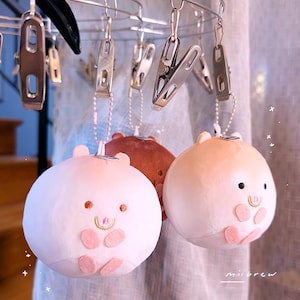 Op de afbeelding: Drie varkensvormige sleutelhangers van pluche in roze en bruine tinten hangen aan een metalen hanger met clips. Elke sleutelhanger heeft een lachend gezicht met kleine ogen en oren en roze accenten. De sleutelhangers hangen voor een licht gordijn.