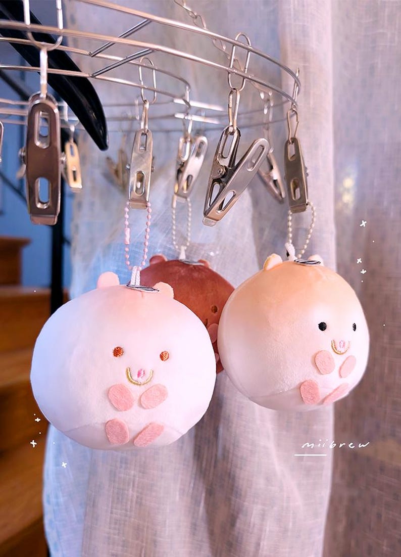 Op de afbeelding: Drie varkensvormige sleutelhangers van pluche in roze en bruine tinten hangen aan een metalen hanger met clips. Elke sleutelhanger heeft een lachend gezicht met kleine ogen en oren en roze accenten. De sleutelhangers hangen voor een licht gordijn.