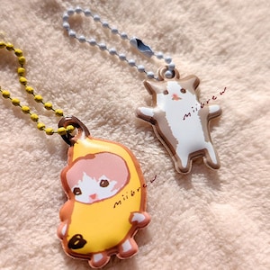 May include: Two keychains featuring cartoon cats. One keychain has a white cat wearing a yellow banana costume with the text "miiibrew". The other keychain has a brown and white cat with the text "miiibrew".