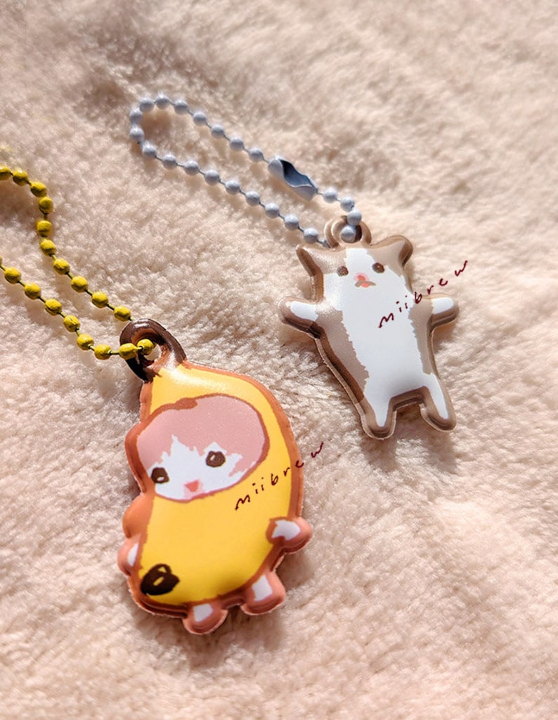 May include: Two keychains featuring cartoon cats. One keychain has a white cat wearing a yellow banana costume with the text "miiibrew". The other keychain has a brown and white cat with the text "miiibrew".
