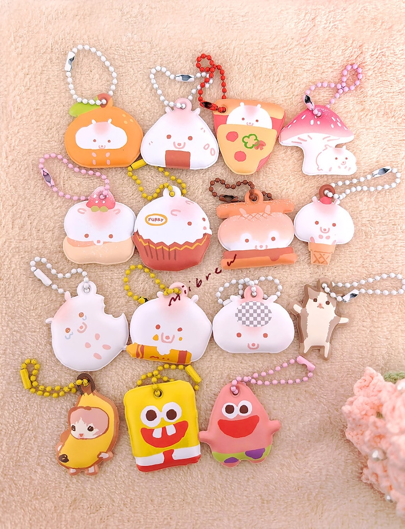 May include: A collection of 14 colorful keychains featuring various cartoon characters and food designs. The keychains are made of soft, plush material and have a variety of colors, including pink, yellow, blue, green, and brown. The keychains are attached to silver chains.