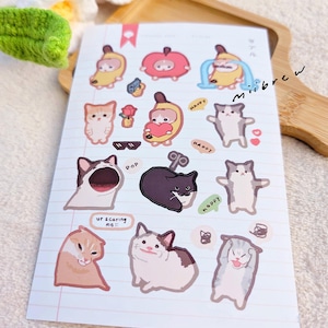 May include: A sheet of stickers featuring various cat illustrations. The stickers are on a white background with pink lines. Some of the stickers feature cats wearing costumes, such as a banana, an apple, and a cat with a heart. Other stickers feature cats with different expressions, such as a cat with sunglasses, a cat with a surprised expression, and a cat with a scared expression. The stickers also include text such as "Happy", "Pop", and "Ur scaring me!!!".