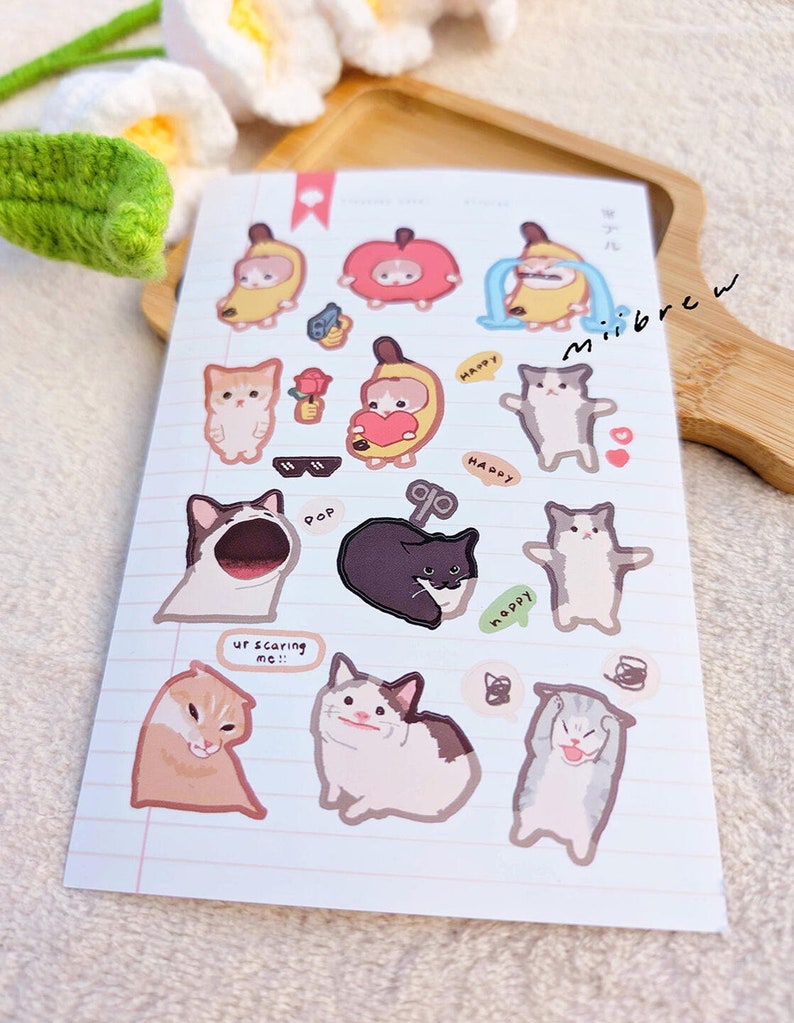 May include: A sheet of stickers featuring various cat illustrations. The stickers are on a white background with pink lines. Some of the stickers feature cats wearing costumes, such as a banana, an apple, and a cat with a heart. Other stickers feature cats with different expressions, such as a cat with sunglasses, a cat with a surprised expression, and a cat with a scared expression. The stickers also include text such as "Happy", "Pop", and "Ur scaring me!!!".