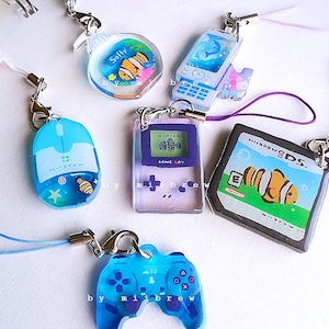May include: A collection of colorful resin charms featuring various designs, including a mouse, a phone, a game controller, and a handheld game console. Each charm has a silver clasp and a matching cord. The charms are decorated with aquatic themes and the text "Softy".