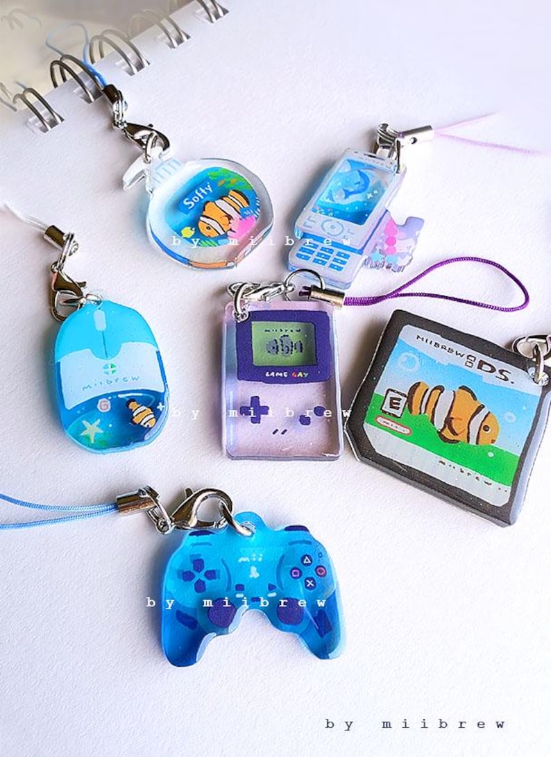 May include: A collection of colorful resin charms featuring various designs, including a mouse, a phone, a game controller, and a handheld game console. Each charm has a silver clasp and a matching cord. The charms are decorated with aquatic themes and the text "Softy".