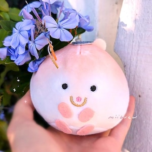 Op de afbeelding: Een pluche perzikvormig speelgoed met een lachend gezicht, zwarte ogen en roze accenten. Het speelgoed is versierd met blauwe bloemen en een kleine bedel. Het woord "miibrew" staat op de zijkant.