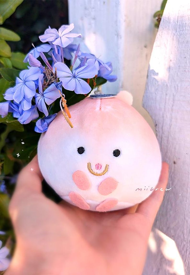 Op de afbeelding: Een pluche perzikvormig speelgoed met een lachend gezicht, zwarte ogen en roze accenten. Het speelgoed is versierd met blauwe bloemen en een kleine bedel. Het woord "miibrew" staat op de zijkant.