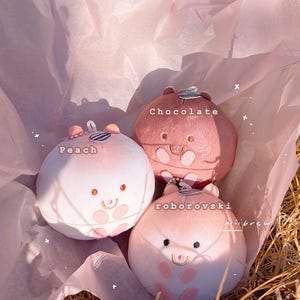Op de afbeelding: Drie ronde, pluche hamster-achtige speeltjes in roze en bruine tinten, met de labels "Peach", "Chocolate" en "roborovski". Elk heeft een klein, lachend gezicht en een gestreept accessoire. Ze liggen op roze vloeipapier.