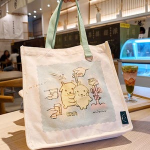 May include: Cream canvas tote bag with pale green handles and a cartoon graphic. The graphic shows two yellow cartoon characters, coffee cups, and the word "WiFi". The bag has a small, dark blue tag with a cartoon face.