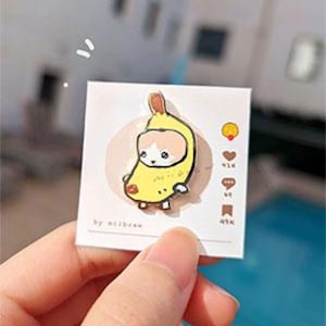 May include: A yellow enamel pin featuring a cartoon cat wearing a banana costume. The pin is on a white card with the text "by milkbrew" and social media icons with numbers.