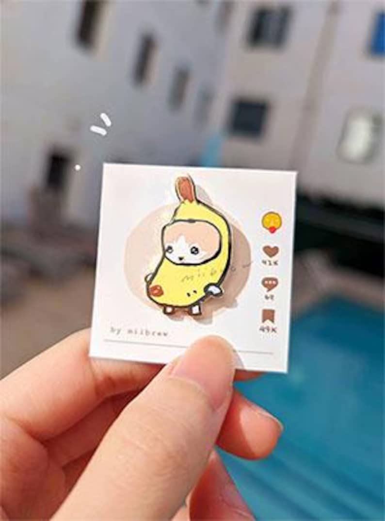 May include: A yellow enamel pin featuring a cartoon cat wearing a banana costume. The pin is on a white card with the text "by milkbrew" and social media icons with numbers.
