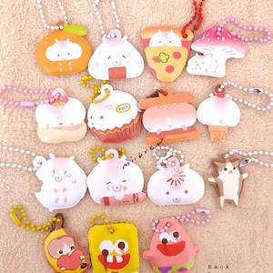 May include: A collection of 14 colorful keychains featuring various cartoon characters and designs. The keychains are made of soft, squishy material and have a variety of shapes, including a mushroom, a pizza slice, a cupcake, a hamburger, and a cat. The keychains are all attached to silver chains.