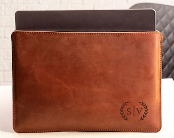 Pode incluir: Uma capa de couro castanho para laptop com um laptop dentro. A capa tem uma borda costurada e um monograma com as letras "S" e "V" dentro de uma coroa de louros. O couro tem um tom rico e quente.