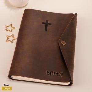 May include: Brown leather journal with a cross and the name "BRIAN" embossed on the front. The journal has a flap closure and a button.