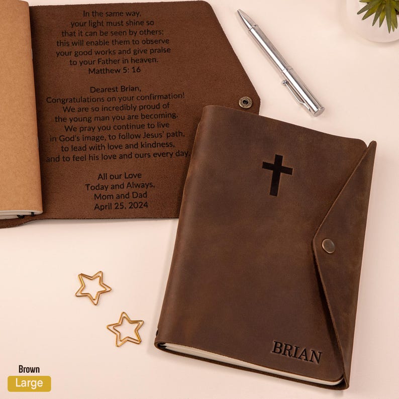 May include: A brown leather journal with a cross symbol and the name "BRIAN" embossed on the cover. The journal is open, revealing handwritten text. Two gold star-shaped paper clips and a silver pen are also visible.