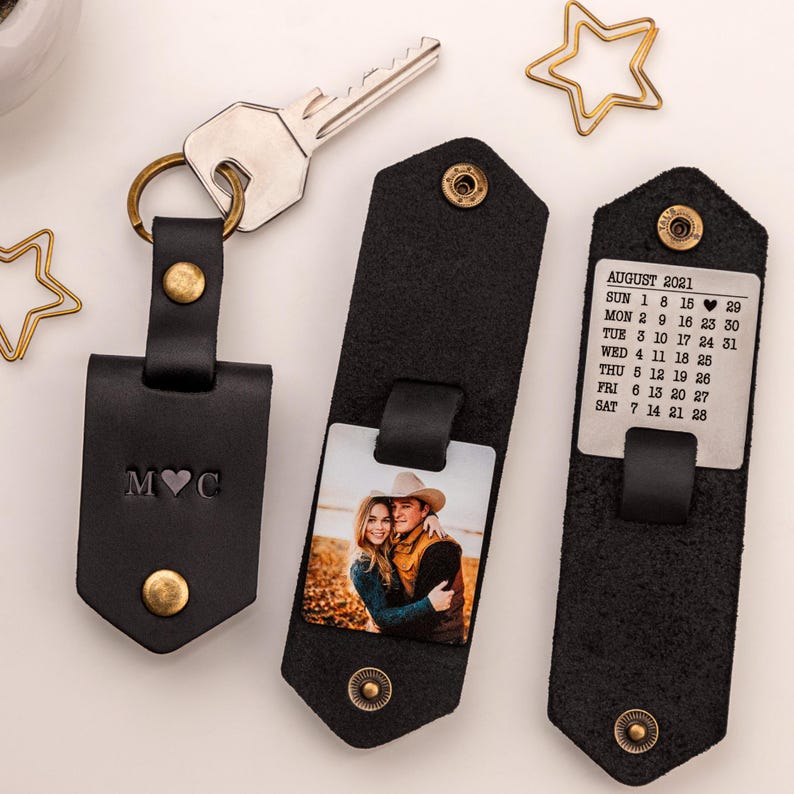Pode incluir: Um chaveiro de couro preto com um inserto para fotos, um inserto de calend&aacute;rio e um charme inicial personalizado. O inserto de fotos mostra um casal em um campo. O inserto de calend&aacute;rio mostra o m&ecirc;s de agosto de 2021. O charme inicial diz "MVC".