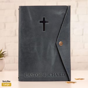 May include: Dusty blue leather Bible cover with a cross cutout and the name "PASTOR RICHARD" embossed at the bottom. The cover has a flap closure with a brass snap. The word "Large" is printed on a yellow label.