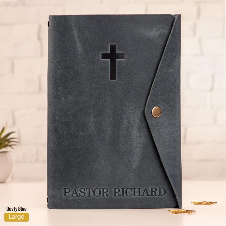 May include: Dusty blue leather Bible cover with a cross cutout and the name "PASTOR RICHARD" embossed at the bottom. The cover has a flap closure with a brass snap. The word "Large" is printed on a yellow label.