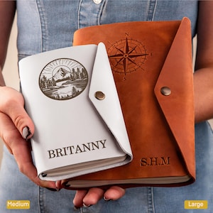 May include: Two leather journals, one white and one brown, are held in the hands. The white journal has a mountain and forest design with the name "BRITANNY". The brown journal features a compass rose design and the initials "S.H.M".