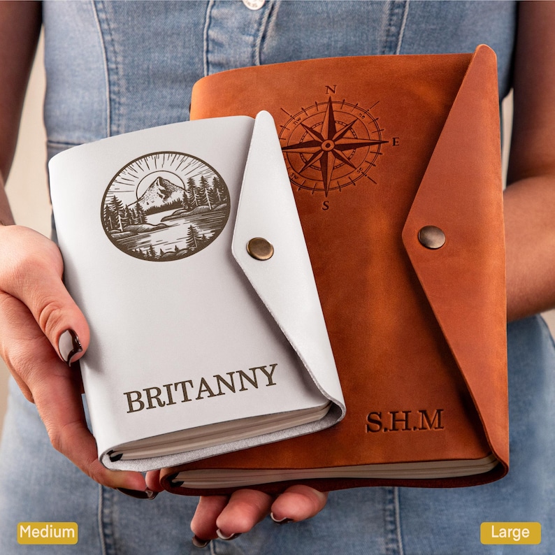 May include: Two leather journals, one white and one brown, are held in the hands. The white journal has a mountain and forest design with the name "BRITANNY". The brown journal features a compass rose design and the initials "S.H.M".