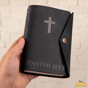 May include: Navy blue leather journal with a cross emblem and the embossed text "PASTOR JEFF". The journal has a flap closure with a brass-colored snap. The journal is held in a person's hand.