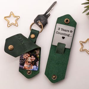 Pode incluir: Chaveiro de foto de couro verde com uma etiqueta de metal prateada que diz "3 Years & Counting..." com um cora&ccedil;&atilde;o. O lado da foto do chaveiro tem uma foto de um casal e seu cachorro. A data "07.18.2021" est&aacute; gravada no couro.