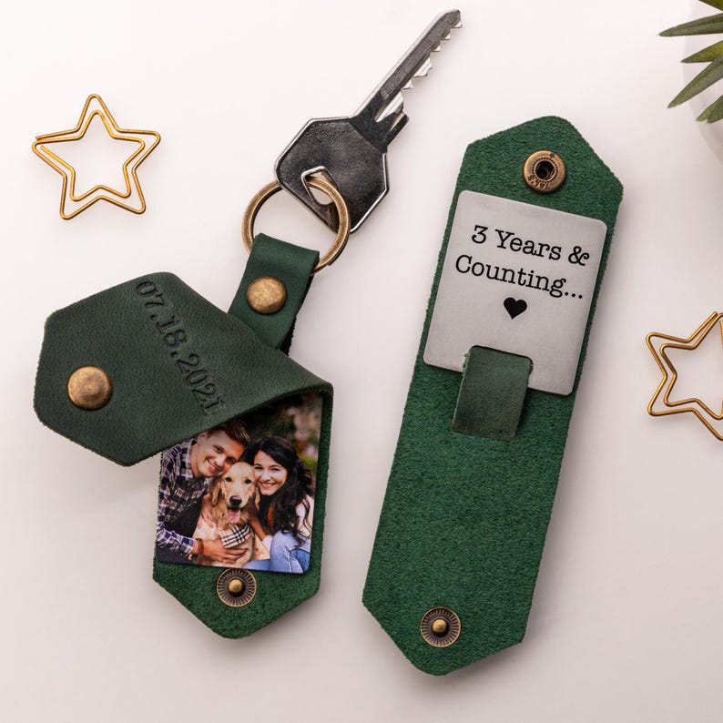 Pode incluir: Chaveiro de foto de couro verde com uma etiqueta de metal prateada que diz "3 Years & Counting..." com um cora&ccedil;&atilde;o. O lado da foto do chaveiro tem uma foto de um casal e seu cachorro. A data "07.18.2021" est&aacute; gravada no couro.