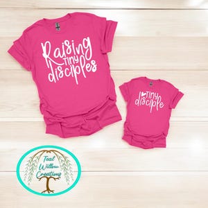 May include: Two pink t-shirts with white text. The larger shirt says "Raising Tiny Disciples". The smaller shirt says "Tiny Disciple".