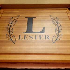 Könnte beinhalten: Ein Schneidebrett aus Holz mit einem schwarzen, gravierten Initial "L" und dem Namen "Lester", umgeben von zwei Lorbeerkränzen.