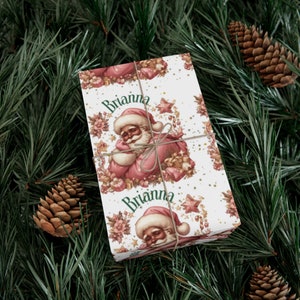 May include: Pink and white Christmas wrapping paper with a repeating pattern of Santa Claus, stars, and candy canes. The paper is personalized with the name "Brianna".