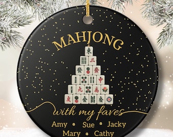 May include: A black round ornament with gold text and accents. The word "MAHJONG" is at the top, with a pyramid of Mahjong tiles below. The text "with my faves" and names are at the bottom. A gold string is attached.