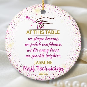 Puede incluir: Adorno redondo de cerámica blanca con una cuerda dorada para colgar. El adorno presenta texto en oro y rosa, incluyendo "AT THIS TABLE" y "JASMINE Nail Technician 2025." También se representa una mano con uñas manicuradas y esmalte de uñas.