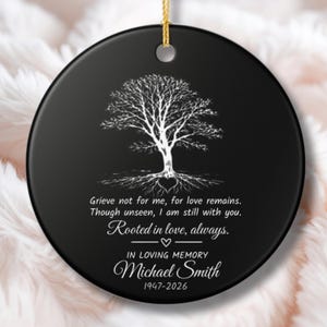 Può includere: Ornamento commemorativo in ceramica nera con un disegno di albero bianco e il testo "Grieve not for me, for love remains." Include il nome Michael Smith e le date 1947-2026. Un filo d'oro è attaccato per appenderlo.
