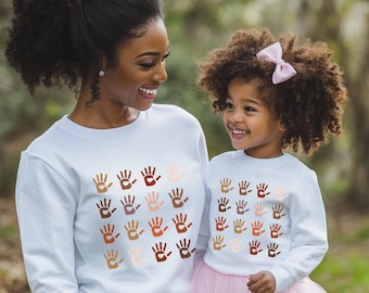 Pode incluir: Uma mãe e uma filha vestindo moletons brancos com um padrão de impressões de mãos coloridas. As impressões de mãos são de diferentes tons de marrom, rosa e bege.