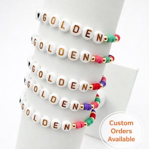 May include: Several white beaded bracelets spelling "GOLDEN" in gold letters. Each bracelet features colourful accent beads in red, pink, green, and purple. Gold-coloured beads separate the letters. The image also includes the text "Custom Orders Available."