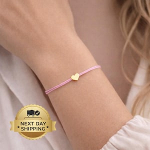May include: A delicate pink bracelet featuring a small gold heart charm. The bracelet is worn on an arm, highlighting the minimalist design. The charm is centered on the bracelet, creating a simple and elegant accessory.