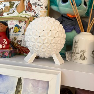 May include: A white geometric sphere with a triangular pattern, supported by a white stand. Other decorative items, including a plush toy and a framed picture, are visible in the background.