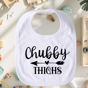 May include: White baby bib with black text that reads "Chubby Thighs" with a black arrow and heart graphic.