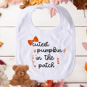 May include: White baby bib with the text "cutest pumpkin in the patch" in black with an orange bow and hearts.
