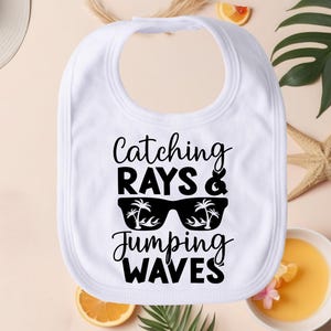 May include: White baby bib with black text that reads "Catching Rays & Jumping Waves" with a pair of sunglasses with palm trees on the lenses.
