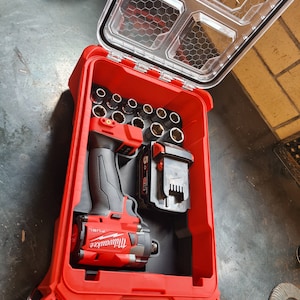 May include: A red Milwaukee Packout tool case with a clear lid, open to reveal a red and black impact wrench, sockets, and a battery. The case is designed for tool storage and organization, with the Milwaukee logo visible.