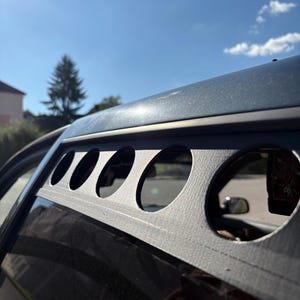 Könnte beinhalten: Nahaufnahme eines schwarzen Autofensters mit einer individuellen Metall-Lüftungsabdeckung. Die Lüftungsabdeckung hat mehrere kreisförmige Aussparungen, die einen Blick in den Innenraum des Autos ermöglichen. Der Himmel ist blau.