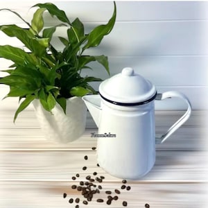 May include: A white enamel coffee pot with a lid, a side handle, and a dark blue rim. The pot features the text FlammSakre printed on the front. Scattered dark brown roasted coffee beans are positioned in front of the coffee pot.