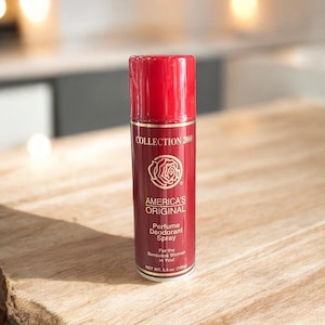 May include: A red and gold perfume deodorant spray bottle. The label reads "COLLECTION 2000 AMERICA'S ORIGINAL Perfume Deodorant Spray". The bottle is on a wooden surface. Net weight is 5.5 oz.
