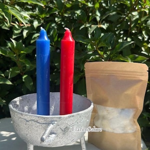 May include: A blue and a red candle stand in a silver cauldron with three legs. A brown paper bag with a clear window is next to the cauldron. The background is green foliage. The text "FlanmSakre" is visible.